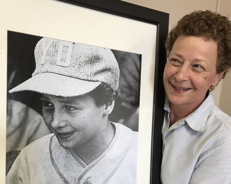 Maria Pepe smiles and holds a large black and white framed photo of herself in her baseball uniform