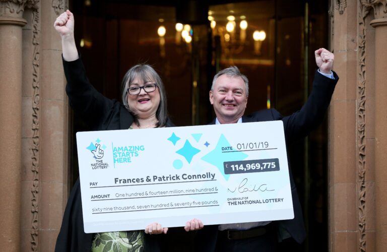 Lottery winner and her husband smile and raise their fists in the air while holding up a giant check