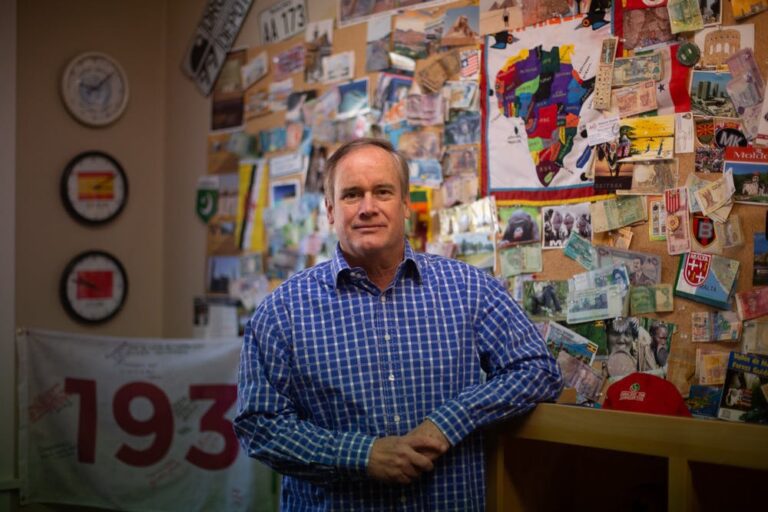 Jim Kitchen smiles in front of a wall filled with bright posters and mementos from his travels