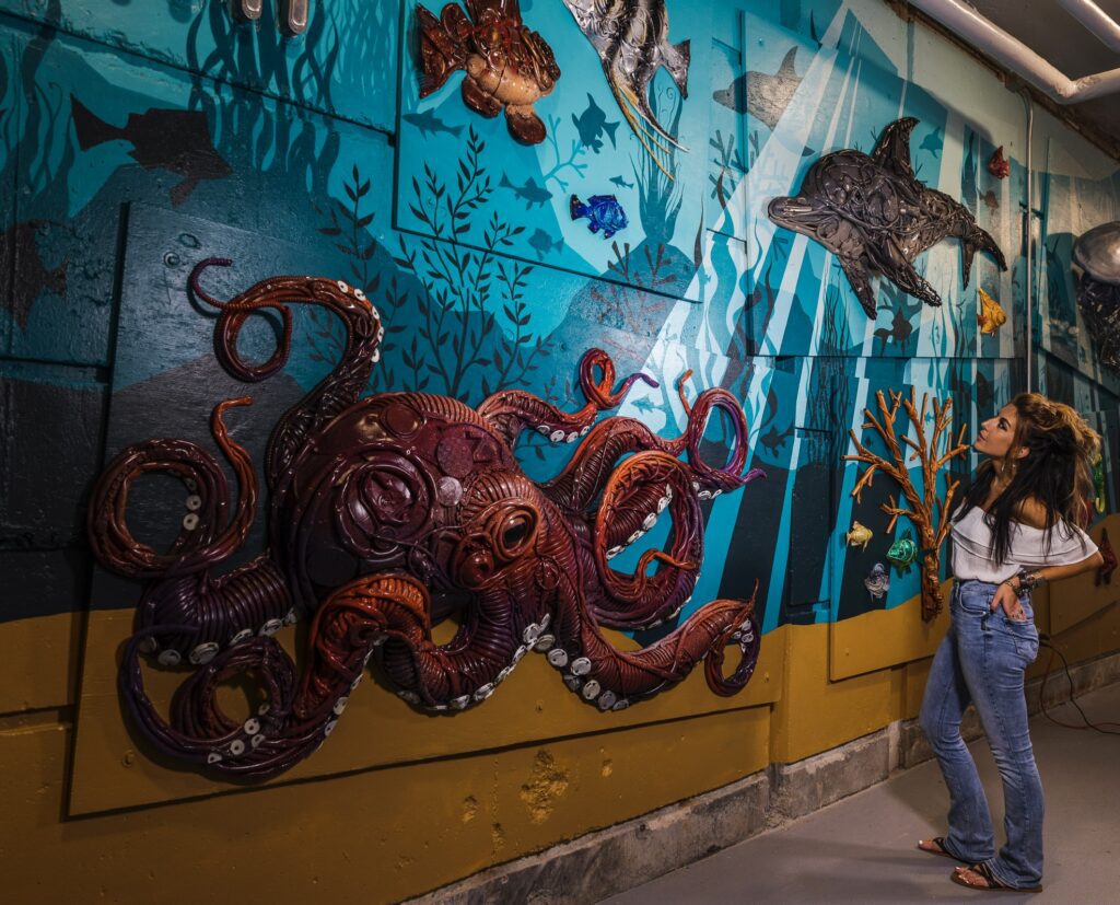Artist Stephanie Hongo stands in front of a giant mural of an underwater scene featuring her sculptures; the focal point is a large octopus