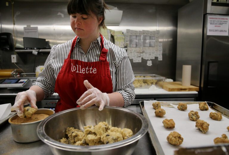 Collette Divitto wears gloves and an apron and stands in front of a big bowl of cookie dough