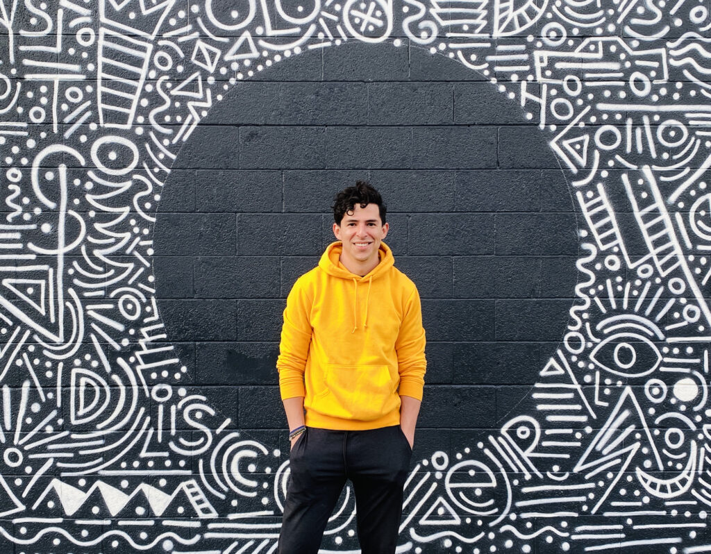 Charlo Walterbach smiles in a yellow sweatshirt in front of one of his murals on a wall