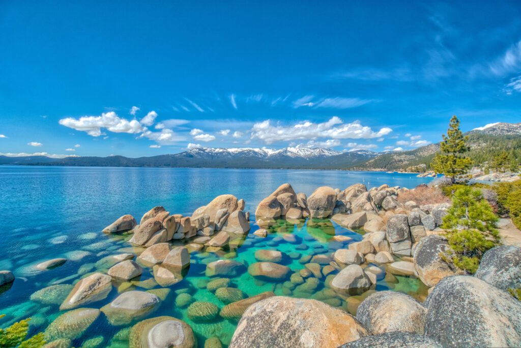 A shot of Lake Tahoe in the winter, stones in the foreground, a clear lake, and mountains in the background under a blue sky