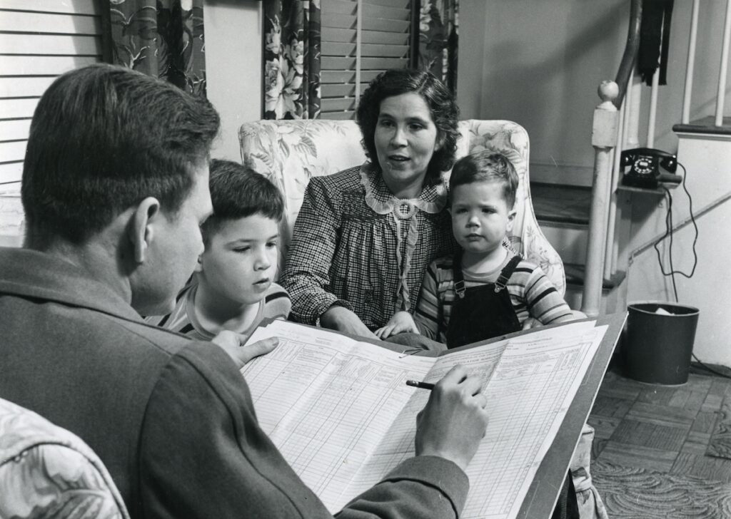 a black-and-white photo of a mother sitting on a chair with her two children while her husband reads the newspaper