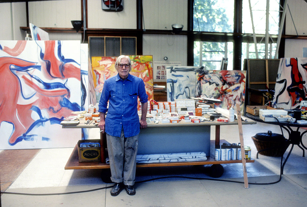 Willem de Kooning stands in front of a workbench in his studio in 1982. He wears glasses and a blue shirt. He has white hair. The room is brightly lit.