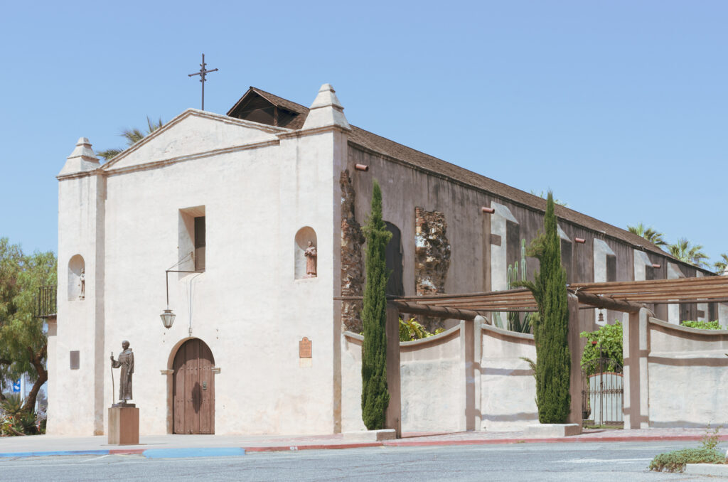 A view of the exterior of Mission San Gabriel, a large white building with a peaked roof and a cross on top