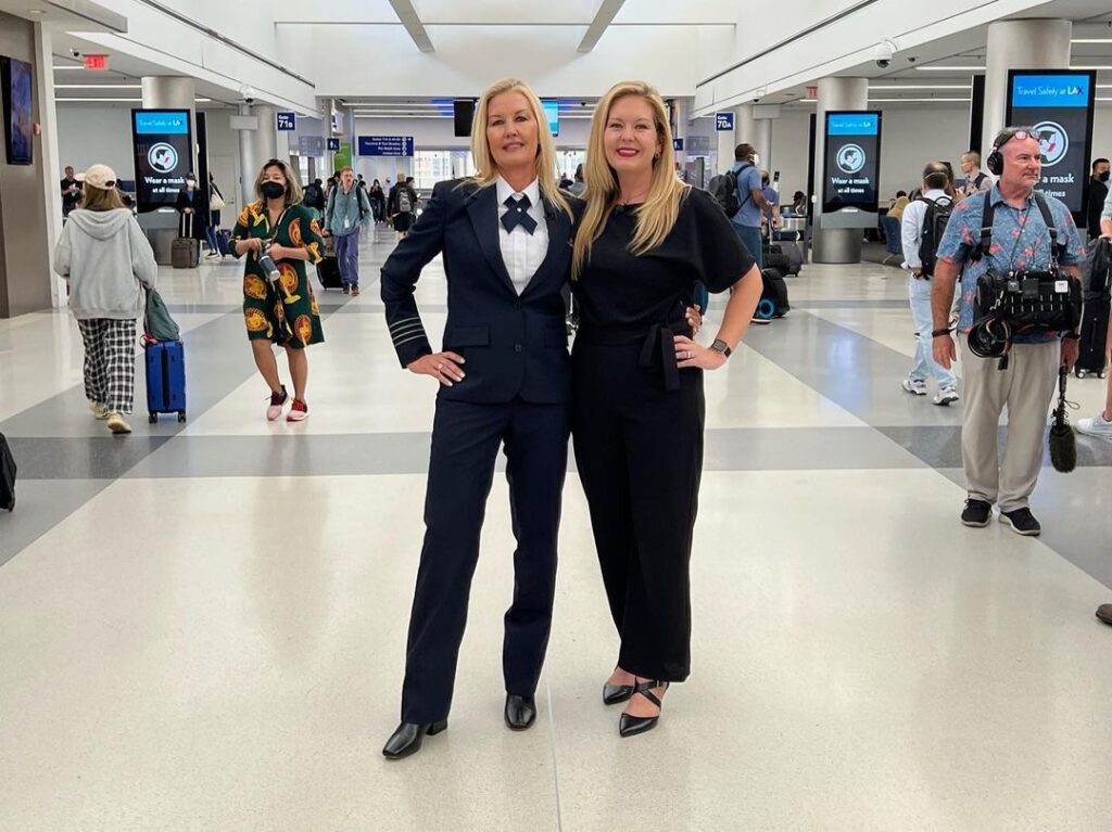 the pilot sisters stand next to each other in the airport smiling with hands on their hips