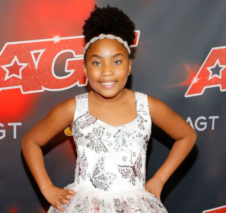 Victory Brinker, the world's youngest female opera singer, stands smiling with hands on her hips in a white dress in front of a red carpet backdrop