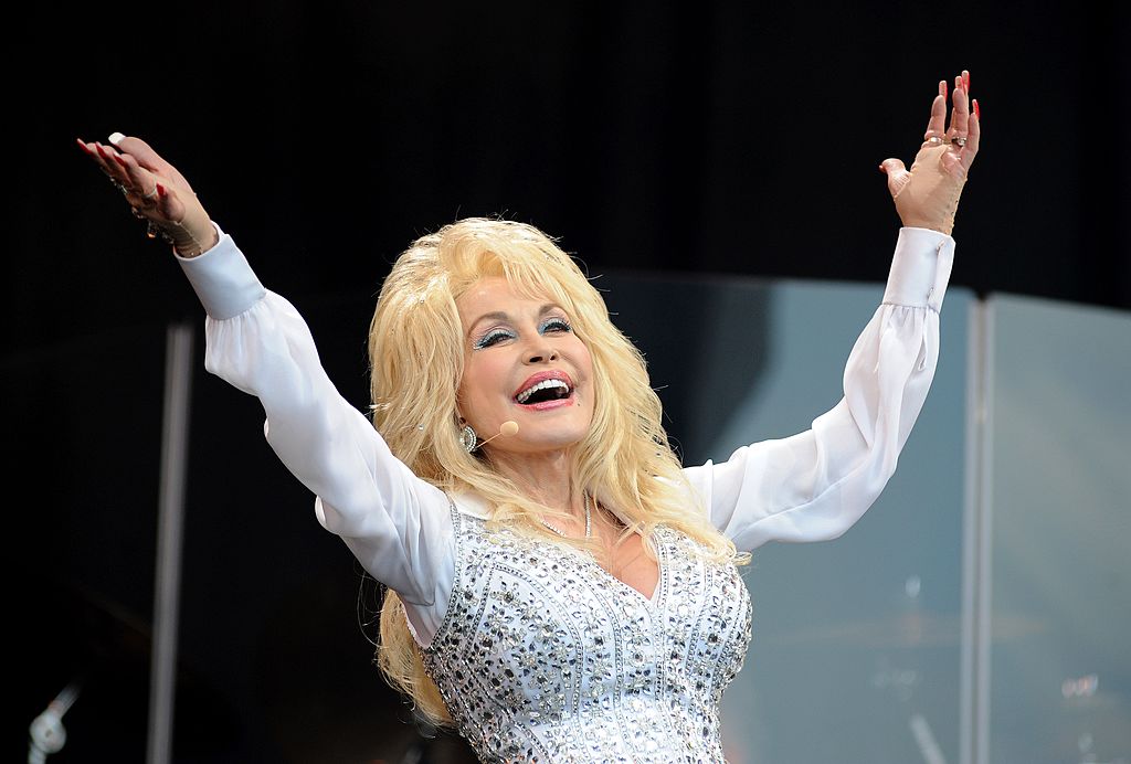 Dolly Parton throws both hands up in the air with a big smile on her face as she performs on stage in 2014