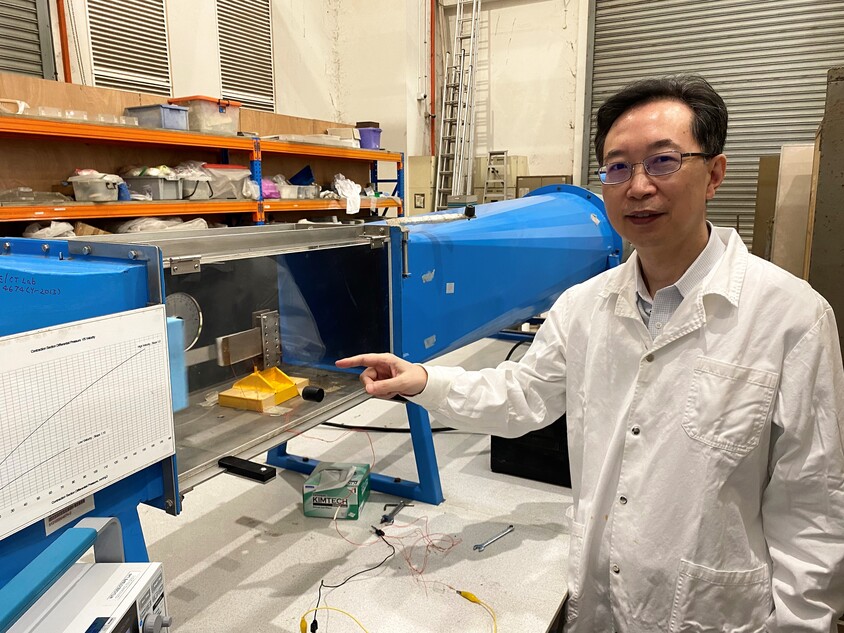 Professor Yang Yaowen stands wearing a white coat in a lab and points to a small wind harvesting device that can turn a gentle breeze into electricity
