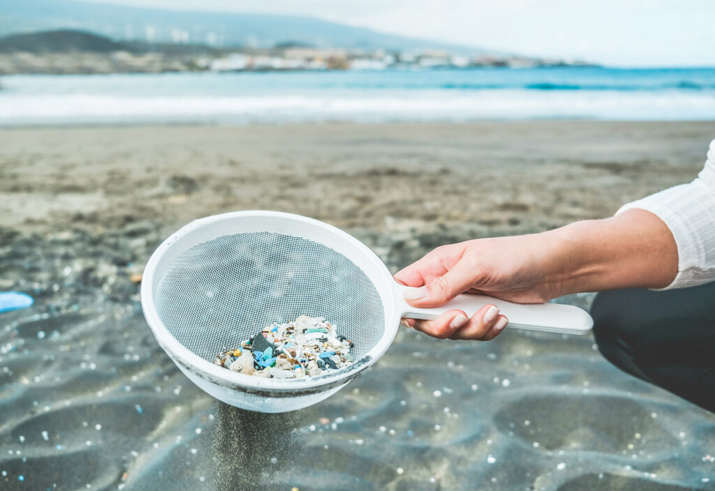 Young woman cleaning microplastics from sand on the beach - Focus on hand