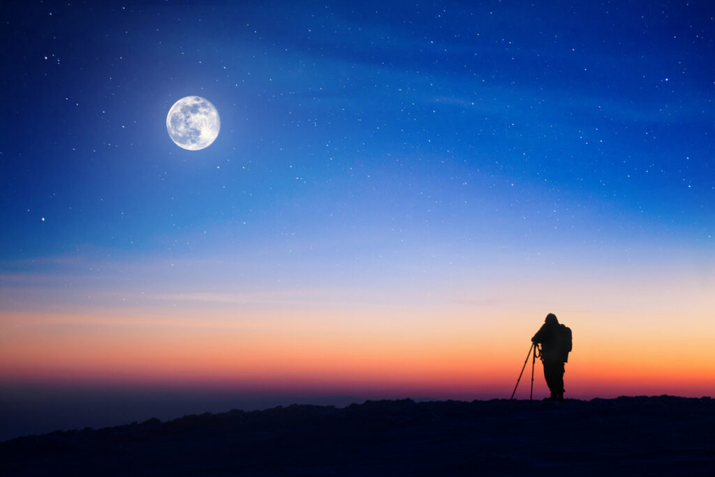 A photographer taking a photo of an moon from the top of a mountain.