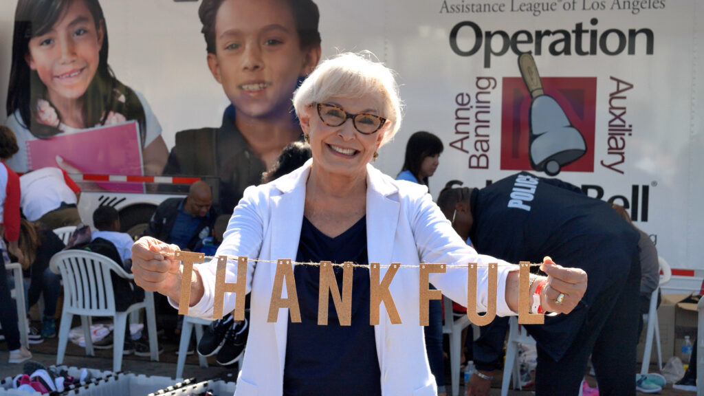 actress Ann Benson stands in front of an Operation School Bell sign and holds up a string of letters that say "Thankful" during filming of the Assistance League documentary A Pebble in the Pond