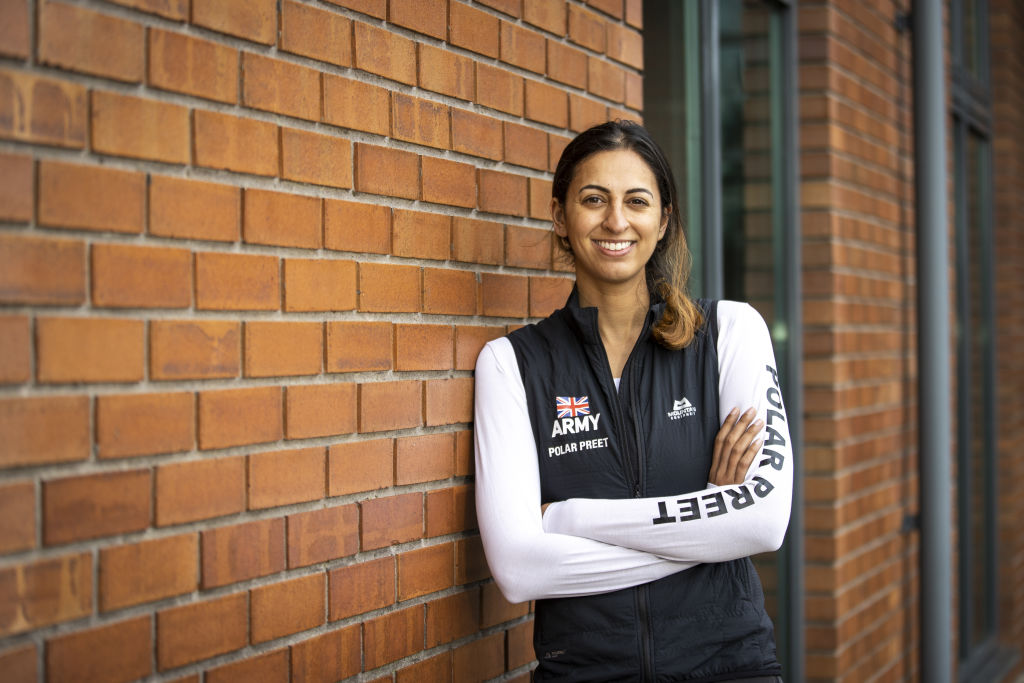 Preet Chandi leans with arms crossed against a brick wall. She's smiling and wearing a shirt that says "Polar Preet" on the sleeve.