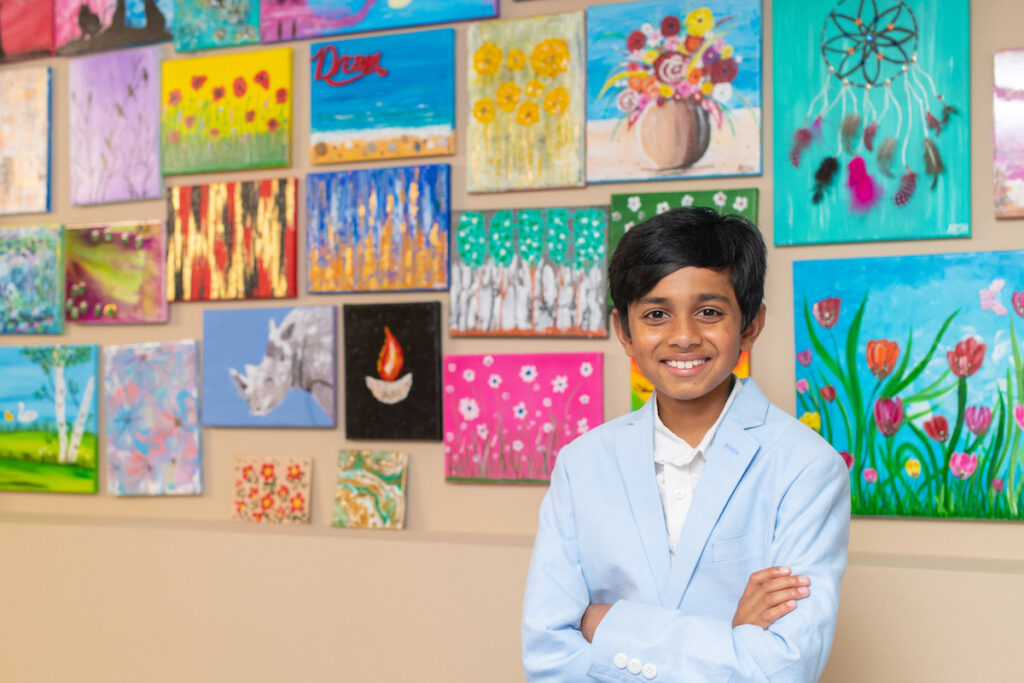 Arsh Pal stands smiling in a blue jacket with arms crossed in front of the paintings he sells for charity