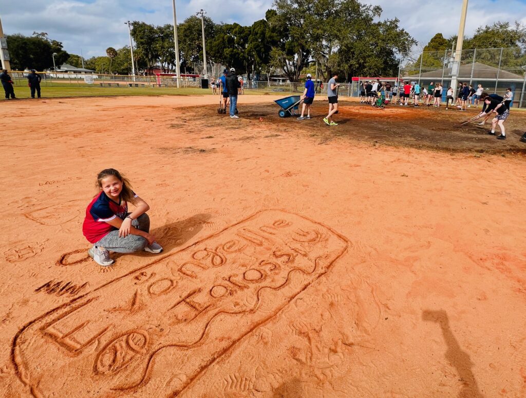 an image of Evangeline Iarossi crouching down on a softball field next to a drawing of her name in the clay