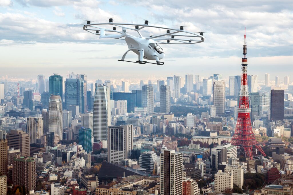 volocopter's electric air taxi, VoloCity, which is white and sleek with an overhead circular propeller, flies over Tokyo in a artistic rendering