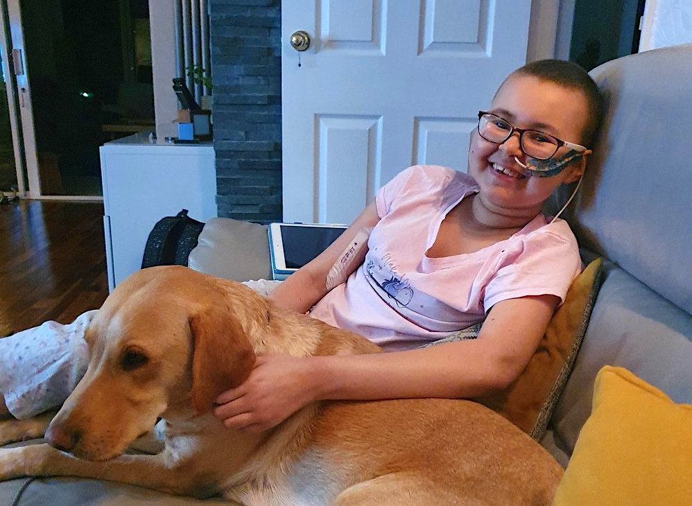 a smiling teenager with glasses, short hair, and a medical device on her face sits with her golden retriever on a couch at home