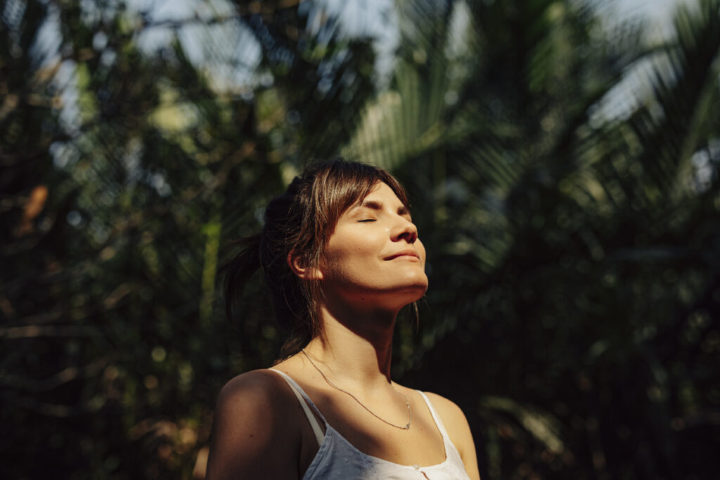 Close up upper body shot of a beautiful happy young woman enjoying the warm sunlight and tropical atmosphere with her eyes closed surrounded by palm trees in a tropical public park.