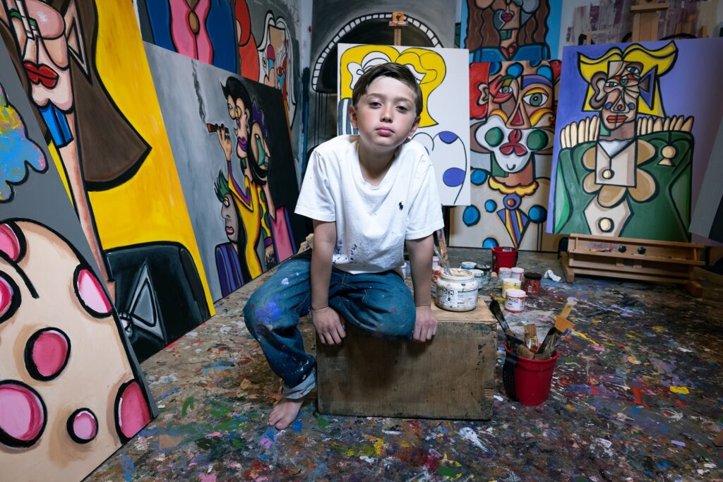 Andres Valencia crouches on his studio in front of a bunch of colorful paintings
