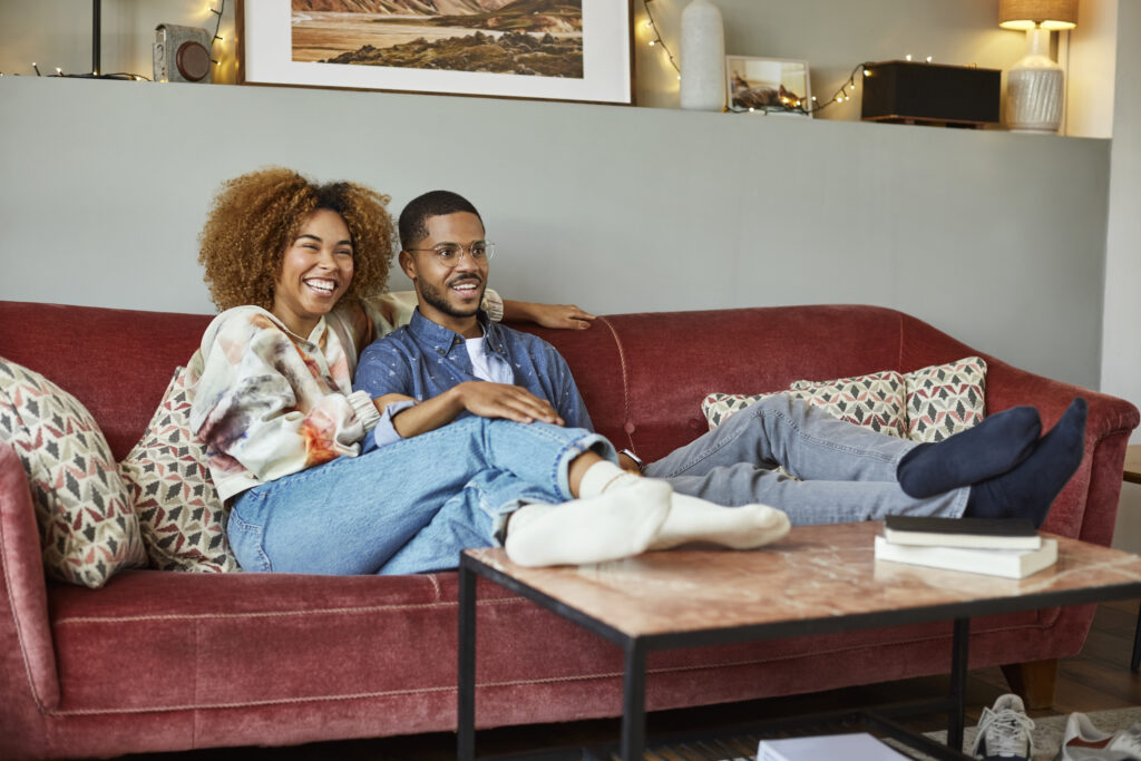 Cheerful woman watching TV with boyfriend while sitting on sofa. Young couple is in living room. They are spending time together at home.