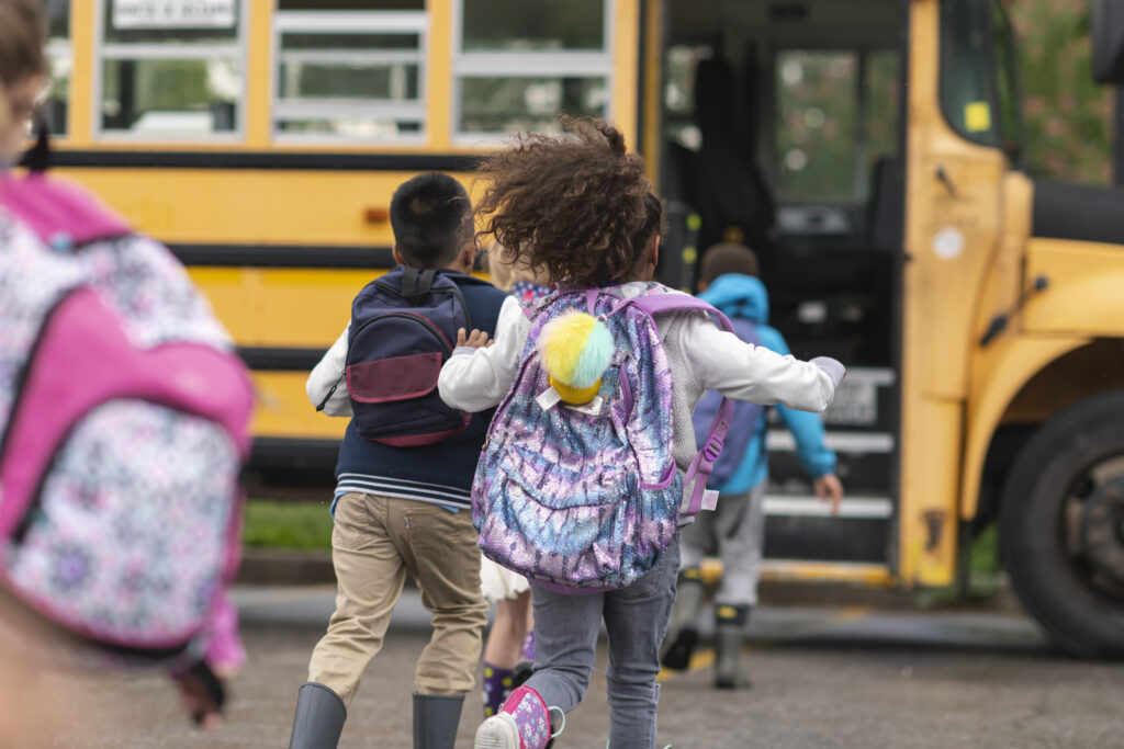 A multi-ethnic group of elementary age children are getting on a school bus. The kids' backs are to the camera. They are running towards the school bus which is parked with its door open. It's a rainy day and the kids are wearing jackets, rain boots and backpacks.