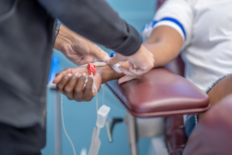 A photo showing a female adult's arm on a chair and a healthcare worker putting tape on the syringe that is on her arm.