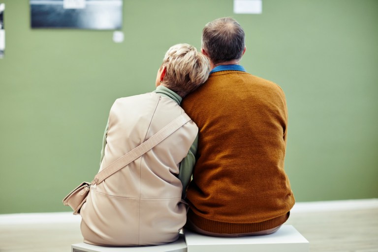 Rear view of unrecognizable mature man and woman in love sitting on stools in modern art gallery looking at artworks