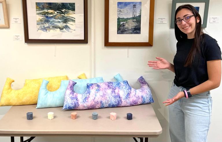 Charlotte Abrams stands next a table featuring her POSH pillow for breast cancer patients in several colors