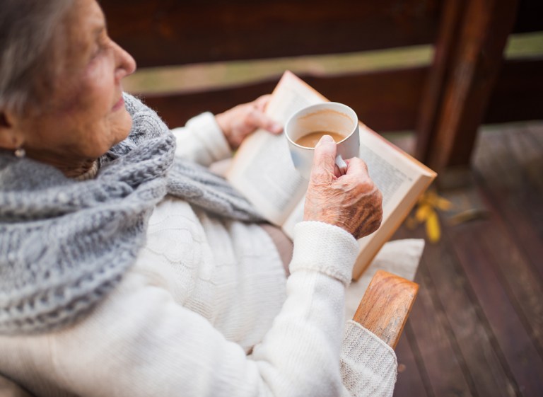 An elderly woman with a cup of coffee or tea sitting outdoors on a terrace on a sunny day in autumn, reading a book.