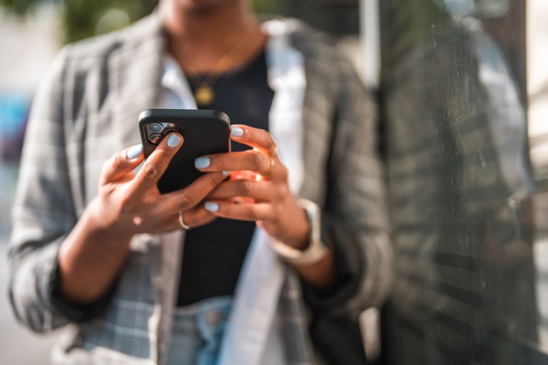 Young African American woman commuting in the city, using a smart phone and texting. She is wearing casual clothes on a sunny day.