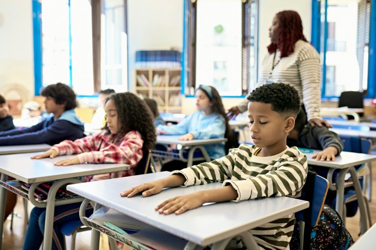 Waist-up view of mature female teacher supporting boys and girls sitting at their child-size desks with arms outstretched and eyes closed.