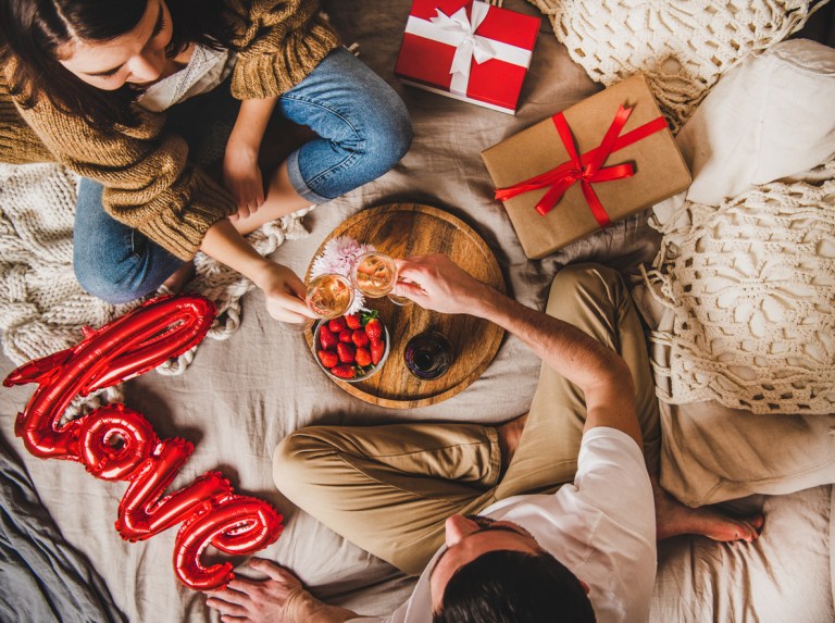 Young happy couple sitting on blankets in bed at home, giving presents and clinking glasses with rose champagne over fruits and a Valentine's Day gift on the bed, top view.