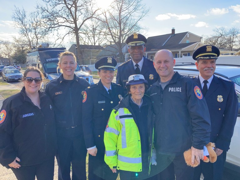 91-year-old crossing guard Louise Kobs wears a bright yellow-green jacket and stands with a group of smiling police officers