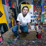 Andres Valencia crouches on his studio in front of a bunch of colorful paintings