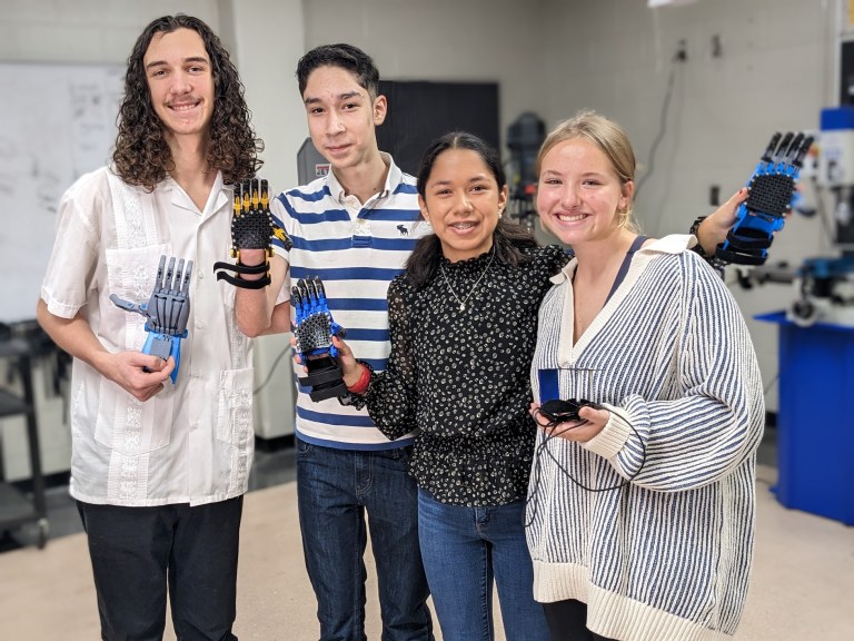 four teenagers smile and group together while holding up robotic hands and parts