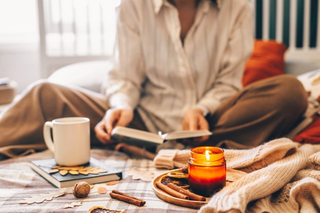 Women sitting on bed reading book surrounded by fall products. Mug coffee or tea and wooden tray with burning candle.